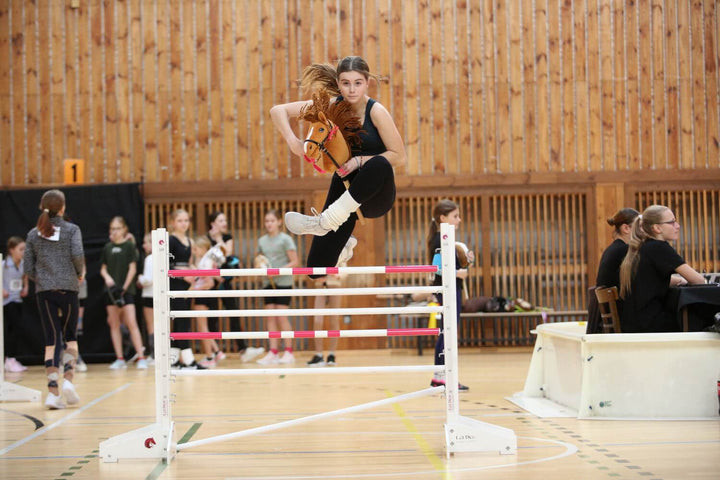 Young participant jumping a pink and white obstacle with a Hobby Horse at an indoor Hobby Horsing event
