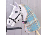 A plush hobby horse by Hobby Horse LarDen, featuring a gray and white face, black bridle, and the Stable Blanket Scottish Cube in blue plaid, stands on a stick before a light wooden wall.
