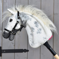 The Hobby Horse LarDen "Hobby Horse Aspen with Black Halter," size S, features a white plush head adorned with black spots, a fluffy mane, and a black halter, all mounted on a wooden stick and displayed against a gray wooden wall.