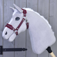 The Halter with fur - leatherette Burgundy by Hobby Horse LarDen, shown on a gray plush hobby horse with white mane and maroon bridle, adds stylish flair against a gray wooden wall—making it the perfect accessory.