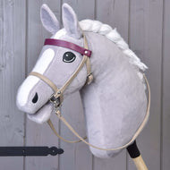 The Hobby Horse LarDen features a gray hobby horse with a white mane, black eye, and a Bordeaux leatherette set—bridle, reins, and martingale—plus a purple browband and cartoon-like face. It stands upright against a wooden background.