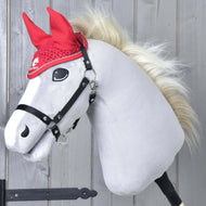 A plush hobby horse from Hobby Horse LarDen, with a white face, black bridle, and blond mane, models the Ear bonnet crocheted Red Sport in front of a light wooden wall.
