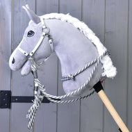 A gray plush hobby horse by Hobby Horse LarDen, featuring the Set Start Silver set (halter, cordeo, lead rope, reins) attached to a wooden stick, displayed against a wooden paneled background.