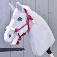 A gray plush hobby horse by Hobby Horse LarDen, featuring a white blaze, black button eyes, and the Rope halter Czi-Czi accessory, is mounted on a wooden stick in front of a gray wooden wall.