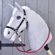 The Reins Czi-czi by Hobby Horse LarDen is a plush grey hobby horse with a white mane, black bridle, and handmade red reins, mounted on a wooden stick and shown against a grey wooden wall.