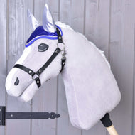 The Hobby Horse LarDen's plush hobby horse features a white mane, black bridle, and the Ear Bonnet DeLuxe Blue for insect protection, all mounted on a wooden stick in front of a light gray wooden wall.