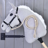 The Hobby Horse LarDen Hobby Horse – Knitted Lead Rope Double Latte, a gray stick horse with black bridle and braided reins, is displayed against a gray wooden wall.