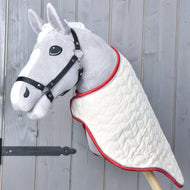 A gray plush hobby horse with a black bridle wears the Hobby Horse LarDen Travel Rug Italy, a quilted white rug with red trim, and is mounted on a wooden pole against a light gray wooden background.