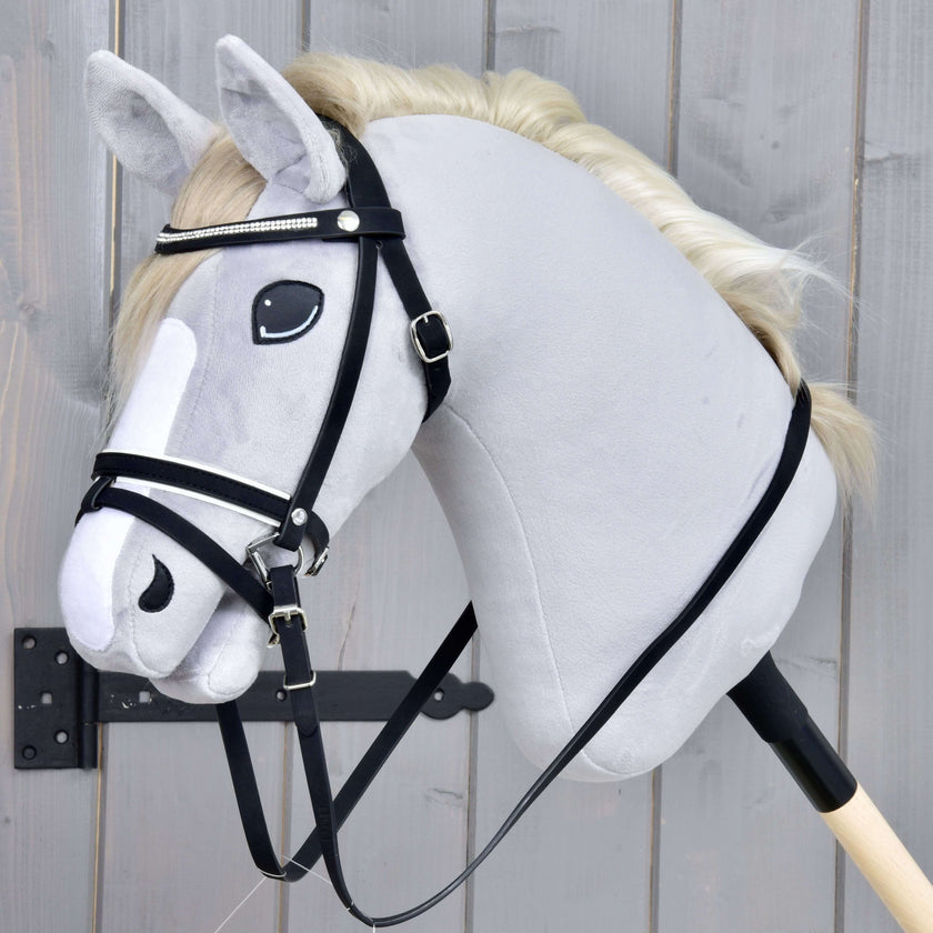 The Hobby Horse LarDen Complete De Luxe bridle with 2 browbands is seen on a realistic gray hobby horse stick toy with a white mane and combined noseband, shown mounted on a wooden stick against a gray wooden background.