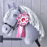 The Hobby Horse Ribbon LarDen Pink by Hobby Horse LarDen is a gray hobby horse with a white yarn mane, black bridle, and a pink and white dressage ribbon on its head, shown against a light wooden background.