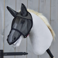 A white hobby horse with a light mane wears the Hobby Horse LarDen Fly Mask De Luxe Full, its face protected by black mesh as it stands against a wooden backdrop.