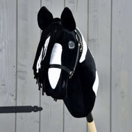 The Hobby Horse Merlin with Black Halter by Hobby Horse LarDen is a black-and-white spotted hobby horse styled after a Barock Pinto stallion, featuring a yarn mane and bridle, mounted on a wooden stick in front of a gray fence.