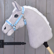 A plush gray stick horse toy with a white mane is shown wearing the Hobby Horse LarDen Halter with fur - leatherette Pastell Blue, propped against a wooden paneled wall with black metal hinges.