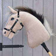 The Hobby Horse LarDen Piccolino with Black Halter, featuring a light tan face and black mane, is mounted on a wooden stick and posed for show jumping in front of a gray wooden wall.