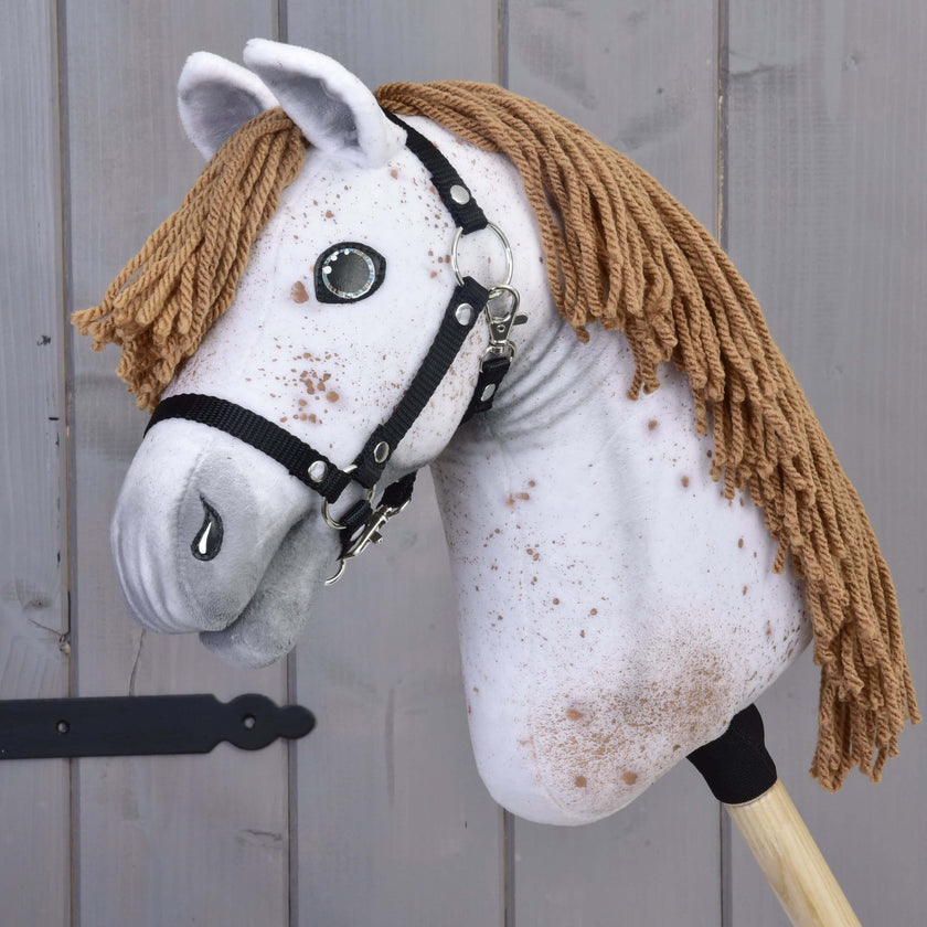 The Hobby Horse LarDen "Hobby Horse Rufus with Black Halter" features a handmade, speckled white fabric head, brown yarn mane, button eyes, and black halter on a wooden stick, shown against a gray wood background.