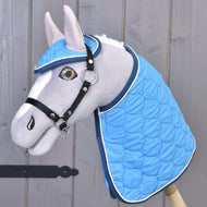 The gray plush hobby horse by Hobby Horse LarDen features the Set Thermo Indigo blanket and matching ear bonnet set with a bridle, displayed as a charming decoration against a wooden wall.