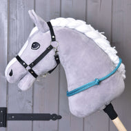 The Cordeo Turquoise by Hobby Horse LarDen is a gray plush hobby horse with a white yarn mane, black bridle, blue rope reins, and a handmade turquoise cordeo for bridleless riding, shown on a stick before a wooden wall.