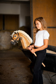 Lifestyle photo: a girl in a white top stands in a stable holding the Hobby Horse Colby with black Halter by Hobby Horse LarDen - palomino plush head, thick cream yarn mane, black halter with silver fittings.