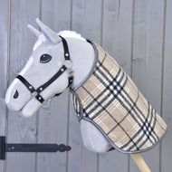 The Hobby Horse LarDen "Blanket Cremo England" is a gray hobby horse with a black halter and plaid beige coat, featuring an ear bonnet for insect protection, mounted on a wooden stick against a gray wooden wall.