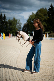 Lifestyle photo: a girl in a black top stands on a sand riding arena under a stormy sky holding the Hobby Horse Frederik LD with black halter by Hobby Horse LarDen, coloured jump poles behind - cream plush head, dark brown yarn mane - shown here with the complete brown bridle with noseband.