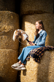 Lifestyle photo: a girl in a denim jacket sits high on round straw bales holding the Hobby Horse Fury with Black Halter by Hobby Horse LarDen - palomino plush head with white markings, cream yarn mane - shown here with the fur-lined LarDen Brown halter and the Bloom Brown lead rope.