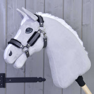 A Halter with fur - leatherette Graphite Full by Hobby Horse LarDen is mounted on a wooden stick and leans against a light gray wooden wall, featuring a gray plush head with white mane, black bridle, and leatherette halter.