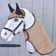 The Set Manchester Le Chocolate by Hobby Horse LarDen features a plush hobby horse with a white face, black harness, brown ears, and textured brown cloth on its head and neck, topped with a Hobby Horse blanket on a wooden stick before a gray wood wall.
