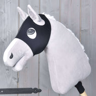 A plush hobby horse with light gray fur, a white mane, and a black bridle wears the Horse Mask without ears Flamingo by Hobby Horse LarDen and is mounted on a wooden stick against a gray wooden panel background.