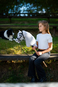 Lifestyle photo: a girl in a white top sits on a garden bench in the evening sun holding the Hobby Horse Popcorn with Black Halter by Hobby Horse LarDen, a border collie resting behind - white plush head, white yarn mane - shown here with the decorated bitless bridle.