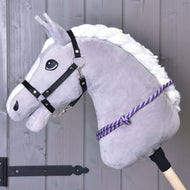 The Hobby Horse LarDen "Cordeo lila stripe" is a gray hobby horse with a white mane, black bridle, and handmade purple polypropylen cordeo on a wooden stick, pictured against a wooden plank wall.