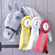 A gray plush hobby horse with white yarn mane displays Hobby Horse LarDen Set Hobby Horse Ribbons NEW—yellow, white, and red ribbons for first, second, and third place—hanging from its neck against a wooden background.