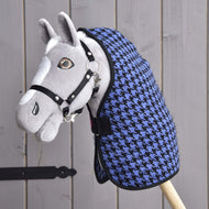 A gray plush hobby horse with a black bridle and the Hobby Horse LarDen Stable Blanket Blue, featuring a blue and black houndstooth pattern, is mounted on a wooden stick and positioned in front of a wooden plank wall.