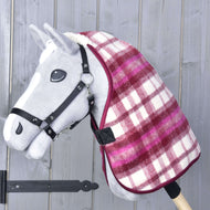 A gray toy hobby horse with a black bridle wears the red and white checked Stable Blanket LarDen by Hobby Horse LarDen over its neck. The background features a wooden wall with vertical planks.
