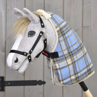 A plush hobby horse with a gray face, white mane, and black bridle wears the Hobby Horse LarDen Stable Blanket Light Blue, is mounted on a wooden stick, and stands before a rustic wooden wall.