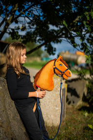 Lifestyle photo: a girl in a black top leans against a stone block under a tree holding the Hobby Horse Wendy with Black Halter by Hobby Horse LarDen - bright chestnut plush head, ginger yarn mane - shown here with the Blues rope stripe lead.