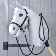 The Hobby Horse LarDen "Western Reins Montana" is a gray plush hobby horse with a white mane and blaze, black bridle with snap hooks, and western reins, mounted on a sturdy wooden stick and displayed on a gray wooden wall.