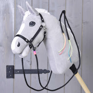 The Hobby Horse LarDen "Split Western Reins Texas" features a gray plush horse with a white mane, black bridle, split western reins, and colorful neck ribbons, mounted on a wooden stick in front of a gray wooden door.