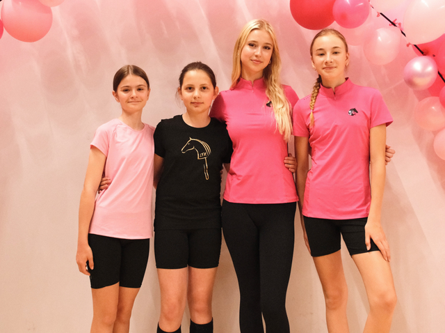 Four young riders stand arm in arm in front of a soft pink backdrop decorated with pink balloons, wearing sporty pink and black tops with black shorts or leggings, including hobby horse logo details. The bright, cheerful setup creates an inviting LarDen