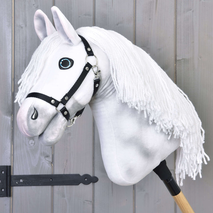 A white plush hobby horse with a black bridle and long yarn mane is mounted on a wooden stick, leaning against a gray wooden fence.
