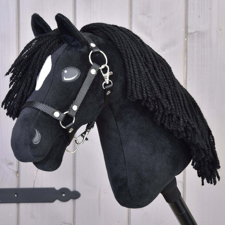 A black plush hobby horse with a white spot on its face, black yarn mane, and a bridle, positioned in front of a light wooden background.