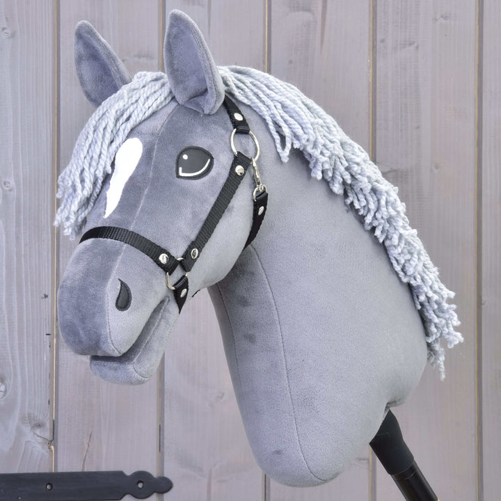 A gray hobby horse with a white spot on its forehead, black bridle, and yarn mane, set against a wooden background.