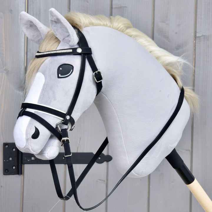 A plush hobby horse with a white mane, black bridle, and a white stripe on its face is attached to a wooden stick, positioned in front of a gray wooden background.