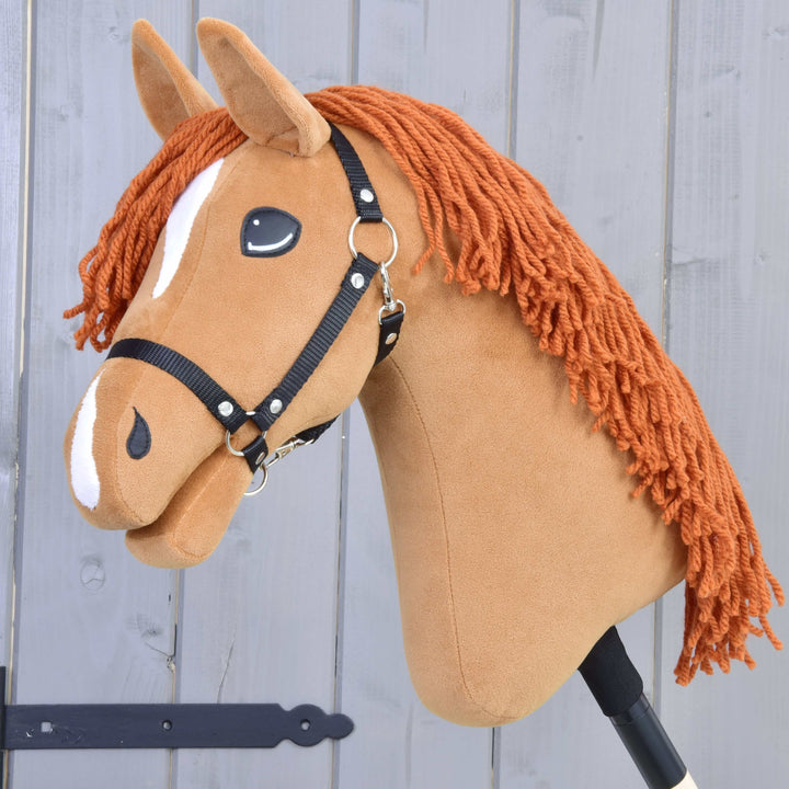 A handmade hobby horse with a brown fabric head, white markings, black bridle, and orange yarn mane, mounted on a stick, in front of a gray wooden background.