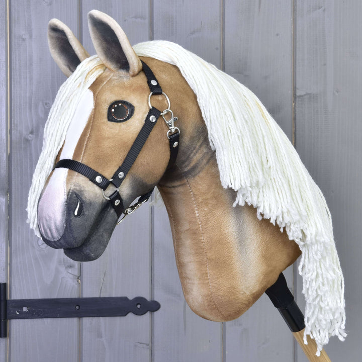 A realistic hobby horse with a tan face, white blaze, black bridle, and long white yarn mane, displayed against a gray wooden wall.