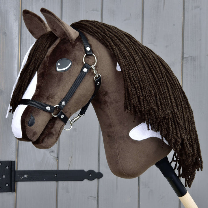 A realistic brown and white hobby horse with a black bridle and long yarn mane, mounted on a wooden stick, is positioned in front of a gray wooden fence.