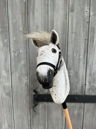 The Hobby Horse LarDen Hobby Horse Luna with Black Halter - size S is shown on a stick in front of a weathered wooden fence with visible grain and a metal hook.