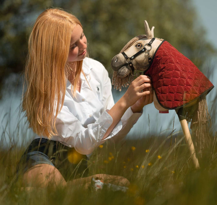 A young person with long blonde hair and a white shirt sits in tall grass, gently adjusting the bridle of a Hobby Horse with a red quilted covering, capturing the imaginative spirit of Hobby Horsing amid blurred greenery in the background.