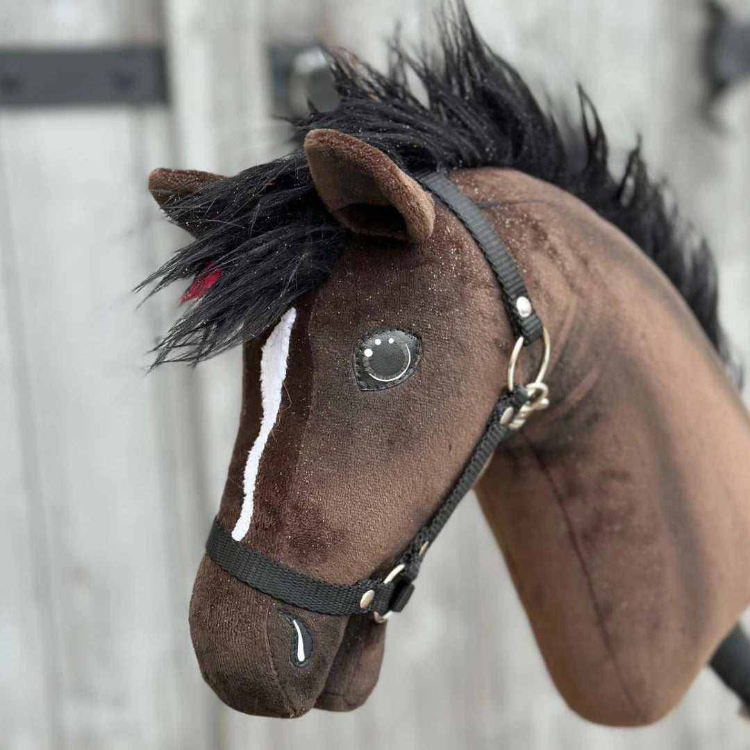 A close-up of Hobby Horse LarDen's "Hobby Horse Lucas with Black Halter," a brown Hannoverian stallion with a black mane, white facial stripe, and black halter, set against a blurred wooden background.