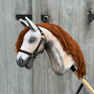 The Hobby Horse Pompo with Black Halter by Hobby Horse LarDen features a gray speckled face, brown yarn mane, and black bridle, inspired by the Hanoverian stallion, and is shown against a weathered wooden fence.