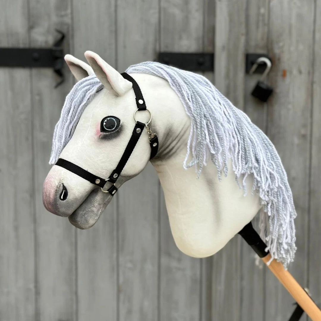 The Hobby Horse LarDen “Hobby Horse Atlas with Black Halter” features a handmade white fabric head, gray muzzle, black halter, and long white yarn mane, shown standing before a weathered wooden door with black metal hinges.
